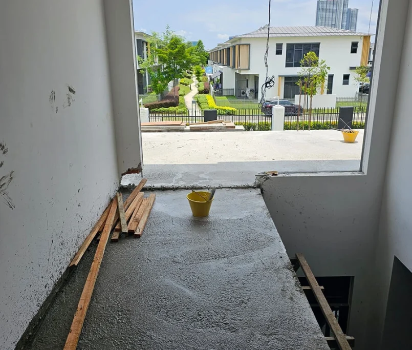 The image captures a clean, newly laid concrete slab extending from the rear of a modern two-storey home. Reinforcement bars indicate further structural additions in progress, while the area below is landscaped with curved walkways, manicured hedges, and young trees. The surrounding upscale neighbourhood and a distant high-rise add context to the residential setting. —Project by Ray & Mist Renovation Sdn. Bhd.