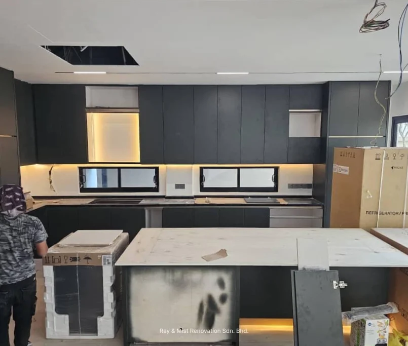 A modern kitchen under renovation featuring dark matte cabinetry, a large kitchen island with a white countertop, under-cabinet lighting, and built-in appliances still in boxes —Project by Ray & Mist Renovation Sdn. Bhd.